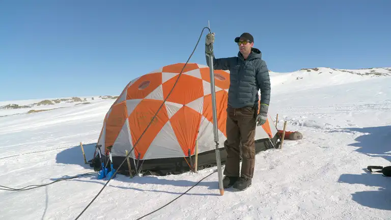 William Colgan st&aring;r foran et telt p&aring; Indlandsisen. Han holder et cirka to meter langt metalsmeltebor i h&aring;nden. Solen skinner.